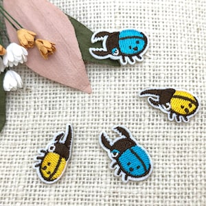 May include: Embroidered patches featuring beetle designs in blue and yellow, with brown accents. The patches are on a textured, off-white surface, with decorative flowers and leaves in the background. These patches are ideal for adding to clothing or accessories.