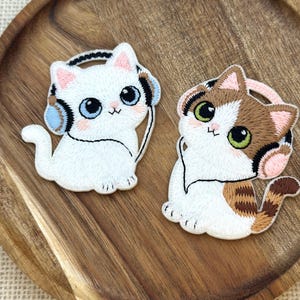 May include: Two embroidered patches featuring cartoon cats wearing headphones. One cat is white with blue accents, the other is calico with brown and tan markings. Both have large eyes and are on a wooden surface.
