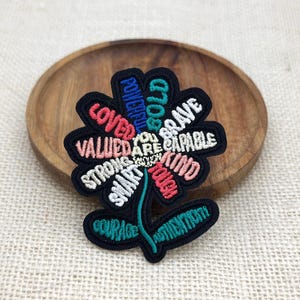 Könnte beinhalten: Ein schwarzer, bestickter Blumenaufnäher mit farbigem Text auf jedem Blütenblatt. Die Wörter umfassen LOVED, POWERFUL, BOLD, BRAVE, CAPABLE, KIND, TOUGH, SMART, STRONG, VALUED und YOU ARE ENOUGH in der Mitte. Stiel und Blätter zeigen die Wörter COURAGE und AUTHENTICITY.