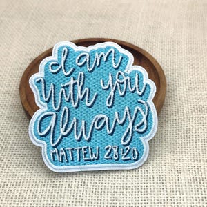 May include: A light blue embroidered patch with the words "I am with you always" in white script. The text "Matthew 28:20" is below the main text. The patch has a white border and is on a wooden surface.