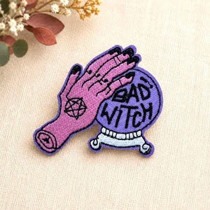Puede incluir: Un parche bordado que muestra una mano rosa con uñas negras y un símbolo de pentagrama negro, sobre una bola de cristal morada con una base azul claro. La bola de cristal contiene el texto bordado en negro BAD WITCH.