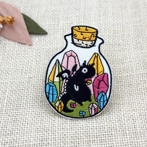 Puede incluir: Parche bordado con un dragón negro dentro de una botella de vidrio con un tapón de corcho. La botella está llena de cristales coloridos en tonos rosa, azul y amarillo. El parche tiene un borde blanco y está sobre un fondo beige.
