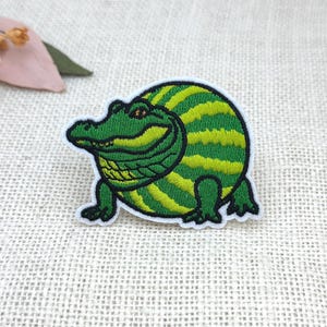 May include: Embroidered patch featuring a smiling alligator with a green and yellow striped body. The alligator has a detailed green head and legs, with a white border. The patch is designed for application on clothing or accessories.