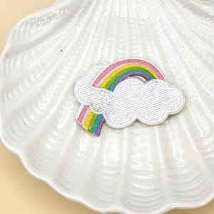 Peut inclure: Un écusson brodé représentant un arc-en-ciel et un nuage. L'arc-en-ciel est composé de rayures rouges, oranges, jaunes, vertes et violettes. Le nuage est blanc avec une texture. L'écusson est posé sur un plat blanc en forme de coquillage.