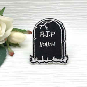 Könnte beinhalten: Schwarz-weißer, gestickter Aufnäher in Form eines Grabsteins. Der Aufnäher trägt in weißen Buchstaben die Aufschrift "R.I.P YOUTH". Eine weiße Rose und grüne Blätter sind im Hintergrund.