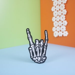 May include: A black and white embroidered patch of a skeleton hand giving the rock and roll hand gesture. The patch is on a light blue surface.