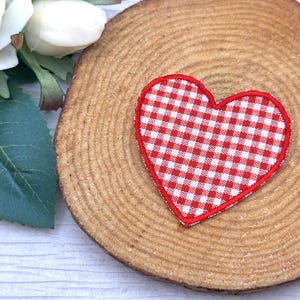 May include: A red and white gingham heart-shaped patch with a red border rests on a wooden slice. White roses and green leaves are in the background. The patch is a decorative element.