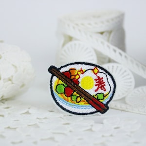 May include: An embroidered iron-on patch featuring a bowl of ramen with chopsticks. The bowl is filled with noodles, vegetables, and a fried egg. The text "素" is embroidered on the egg.
