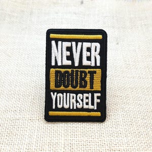 May include: A rectangular embroidered patch with a black background and a gold border. The text NEVER DOUBT YOURSELF is stitched in white and black lettering, with the word DOUBT centered on a gold horizontal bar across the middle of the patch.