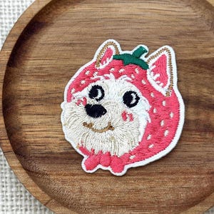 May include: An embroidered patch featuring a white dog with a black nose and eyes, wearing a pink strawberry-shaped hood with a green stem on top. The patch has a white border and detailed stitching, showcasing a playful design of a canine character.