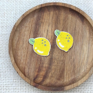 May include: Two small embroidered patches shaped like yellow lemons with green leaves. Each lemon patch features a bright yellow body, a small green leaf at the top, and white highlights to indicate texture and light reflection on the fruit surface.