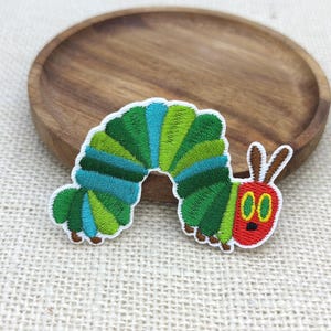 May include: An embroidered patch shaped like a caterpillar with a red face, two brown antennae, and a segmented body featuring alternating shades of green and blue. The design has a white border and is stitched with dense, textured thread throughout.