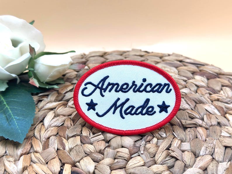 K&ouml;nnte beinhalten: Ein ovaler, bestickter Aufn&auml;her mit wei&szlig;em Hintergrund und einem leuchtend roten Rand. Der Text "American Made" ist in dunkelblauer Schreibschrift gestickt und wird von zwei dunkelblauen, gestickten Sternen links und rechts flankiert.