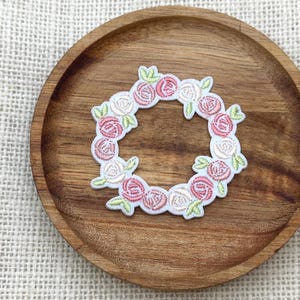 Peut inclure: Un écusson brodé circulaire représentant une couronne de petites roses roses et blanches avec des feuilles vert clair. La composition florale est réalisée avec un travail de fil détaillé, créant un motif circulaire délicat et texturé.