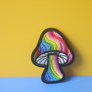 Może przedstawiać: Naszywka z haftowanym tęczowym grzybem z czarnym obramowaniem. Grzyb znajduje się na żółtym i niebieskim tle.