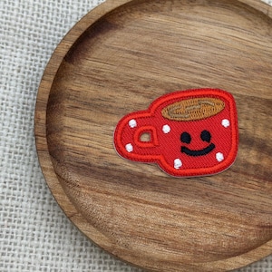 May include: A red embroidered patch in the shape of a coffee cup with a smiling face and white polka dots. The cup has a brown coffee fill and is set on a wooden plate.