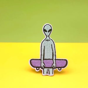 May include: Embroidered patch featuring a gray alien figure holding a purple skateboard. The alien has large black eyes and a small bow tie. The patch is outlined in black and set against a yellow and green background.