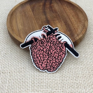 May include: An embroidered patch featuring a pink human brain being knitted with black needles by two pale hands. The design is outlined in white thread, showcasing detailed anatomical folds of the brain and the motion of knitting needles.