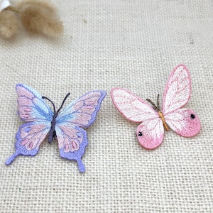May include: Two embroidered butterfly patches. The left patch features a gradient of purple, blue, and light pink thread with dark antennae. The right patch is primarily light pink with darker pink accents, a brown body, and small black spots on the wings.
