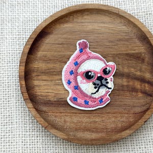 May include: Embroidered patch featuring a cartoon seal wearing a pink hat and sunglasses. The seal's hat has blue star-shaped accents. The patch is placed on a round wooden tray, perfect for adding to clothing or accessories.