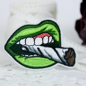 May include: Green embroidered patch of a mouth with white teeth biting down on a white and brown cigarette.