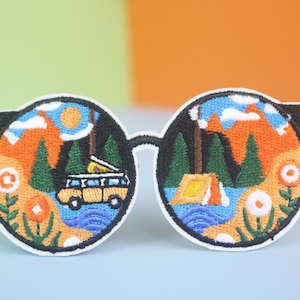 May include: Embroidered patch of a pair of round sunglasses with a view of a mountain landscape, a yellow camper van, and a tent. The sunglasses are white with black frames.