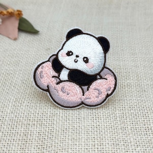 Op de afbeelding: Geborduurde patch met een schattige panda beer die op een roze wolk zit. De panda heeft zwart-witte vacht met roze wangen. De wolk is zachtroze met een grijze omtrek. Voor kleding of accessoires.