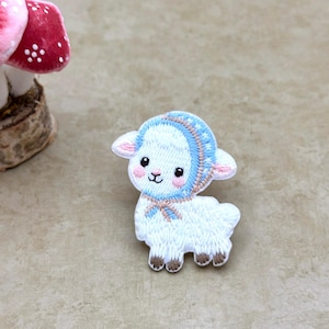 Pode incluir: Patch bordado com um cordeiro branco usando um gorro azul e um laço a condizer. O cordeiro tem orelhas e bochechas cor-de-rosa e cascos castanhos. Um cogumelo caprichoso com manchas vermelhas e brancas está no fundo.
