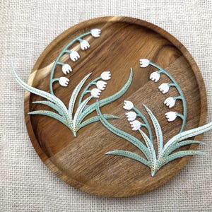Puede incluir: Dos parches bordados de lirios de los valles con flores blancas en forma de campana y hojas de color verde salvia claro con detalles en hilo metálico dorado. Los motivos florales muestran tallos curvos sobre una bandeja de madera redonda.