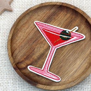Pode incluir: Um emblema bordado em forma de taça de martini com contorno rosa e preenchimento vermelho. A taça contém uma azeitona verde-escura num palito de madeira. O emblema tem uma borda branca e está exposto sobre uma bandeja de madeira circular.