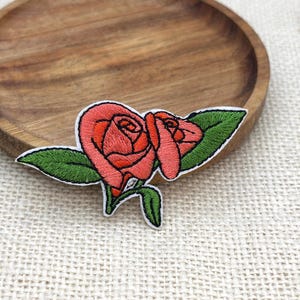 Puede incluir: Un parche bordado que presenta dos rosas de color rosa coral con costuras detalladas, acompañadas de tres hojas de color verde vibrante y un pequeño tallo verde. El diseño está delineado con hilo blanco, creando un borde claro alrededor.
