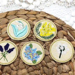 May include: Five embroidered floral patches with gold trim. Designs include lavender, blue hydrangeas, white flowers, yellow flowers, and blue flowers. The patches are on a woven background.