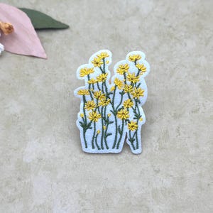 May include: Embroidered patch featuring a cluster of yellow flowers with green stems and leaves. The patch is white with a floral design. The patch is approximately 2 inches tall and 1.5 inches wide.