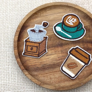 Pode incluir: Três emblemas bordados com temas de café: um moinho de café de madeira castanha com topo prateado, uma chávena e pires azul-petróleo com latte art e uma colher branca, e um copo de café bege com tampa castanha e um grão de café.