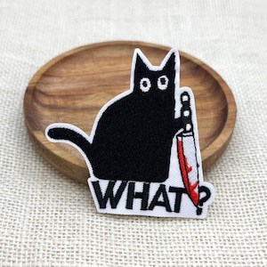 May include: Black cat patch holding a bloody knife with the word "WHAT?" in white block letters. The cat has large, wide eyes and a curved tail. The patch is on a wooden plate.