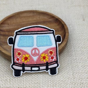 Può includere: Una toppa ricamata a forma di furgoncino vintage con esterno rosa e bianco, finestrini azzurri e bordi neri. La parte anteriore presenta un emblema centrale con il simbolo della pace e diversi disegni di fiori gialli e rossi sul pannello inferiore.