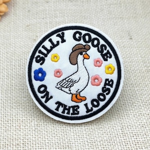 Puede incluir: Un parche bordado circular con fondo blanco y borde negro. En el centro, una oca blanca lleva un sombrero de vaquero marrón y tiene patas naranjas. Pequeñas flores amarillas, rosas y azules rodean a la oca. El texto SILLY GOOSE ON THE LOOSE está bordado en letras mayúsculas negras alrededor del borde.