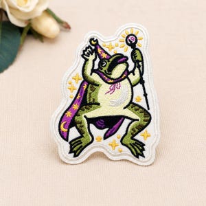 May include: An embroidered patch featuring a green frog dressed as a wizard, wearing a purple cape and hat with yellow stars and moons. The frog holds a staff topped with a glowing orb, surrounded by small yellow stars on a white background.