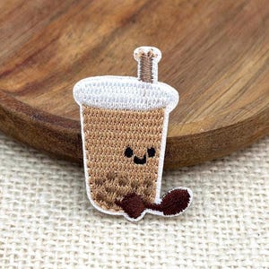 Könnte beinhalten: Ein gestickter Aufnäher in Form eines Bubble-Tea-Bechers mit hellbrauner Flüssigkeit, weißem Schaum oben und einem braunen Strohhalm. Der Becher zeigt ein einfaches, schwarz gesticktes Gesicht mit zwei Augen und einem Lächeln sowie dunkelbraune Perlen am Boden.