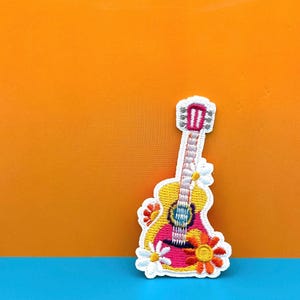 Puede incluir: Un parche bordado blanco de una guitarra rosa y amarilla con margaritas blancas y flores naranjas.