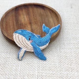 May include: An embroidered patch featuring a blue whale with a light tan underbelly and textured stitching. The whale is depicted in a swimming position with its tail fin raised and a small pink detail near the eye, showcasing detailed thread work.