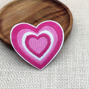 May include: Embroidered heart patch in shades of pink and white. The patch features a layered design with a white border. The heart is placed on a textured, neutral-coloured surface, with a wooden plate in the background.