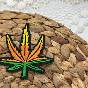 May include: Embroidered patch in the shape of a cannabis leaf. The leaf features a black outline with orange, yellow, and green stitching. The patch is placed on a woven, brown, circular surface.