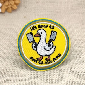 May include: A circular embroidered patch featuring a white goose with orange feet and beak, holding two silver meat cleavers. The background is bright yellow with a blue and gold border. The text reads, It's okay to feel all the feels.