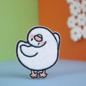 May include: White embroidered patch of a small bird with black eyes and a pink blush. The bird is standing on two legs and has its wings folded.
