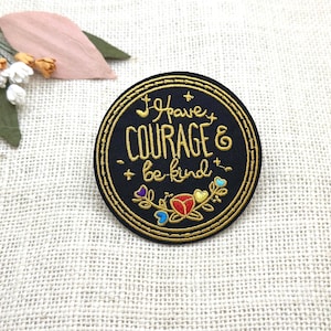 Op de afbeelding: Een ronde, zwarte geborduurde patch met gouden letters met de tekst "I have COURAGE & be kind". De patch heeft een gouden rand en kleurrijke bloemenaccenten. Een klein boeket bloemen en bladeren bevindt zich in de linkerbovenhoek.