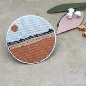 May include: Circular embroidered patch with a landscape design, featuring a sun, horizon, and layered earth tones. The patch has a white border. The design incorporates shades of orange, brown, and grey. Floral decorations are in the background.