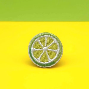 May include: Embroidered patch in the shape of a lime slice. The patch features a green rind with white and light green segments. The patch is set against a bright yellow background with a green upper section.