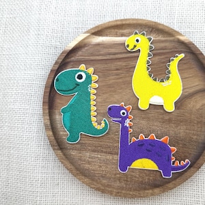 Puede incluir: Tres parches de dinosaurios coloridos sobre una placa de madera. Los parches son verdes, amarillos y morados, cada uno con un diseño único. El dinosaurio verde está de pie, el amarillo es alto y el morado tiene un cuello largo. La placa está sobre una superficie texturizada blanquecina.