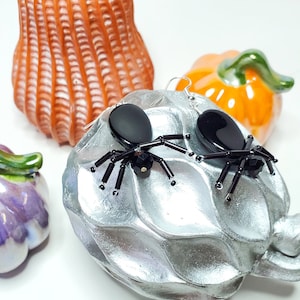 以下が含まれることがあります： 黒いビーズのアクセントが付いた黒いクモのイヤリングのペア。イヤリングは、小さなカボチャを背景にした銀色の背景の上にあります。